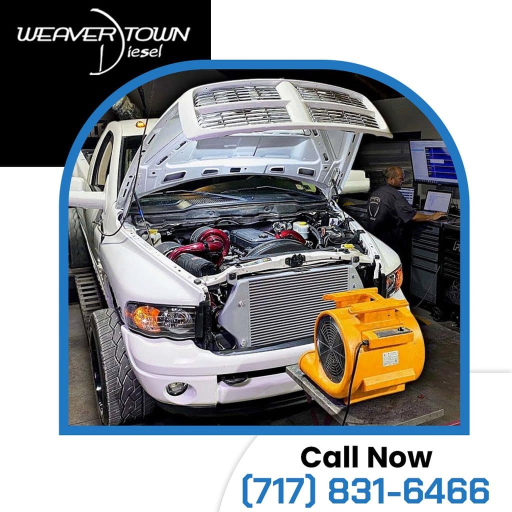 A white Ram diesel truck strapped to a chassis dyno with cooling fans in front of the intercooler while a technician monitors performance data on a computer.