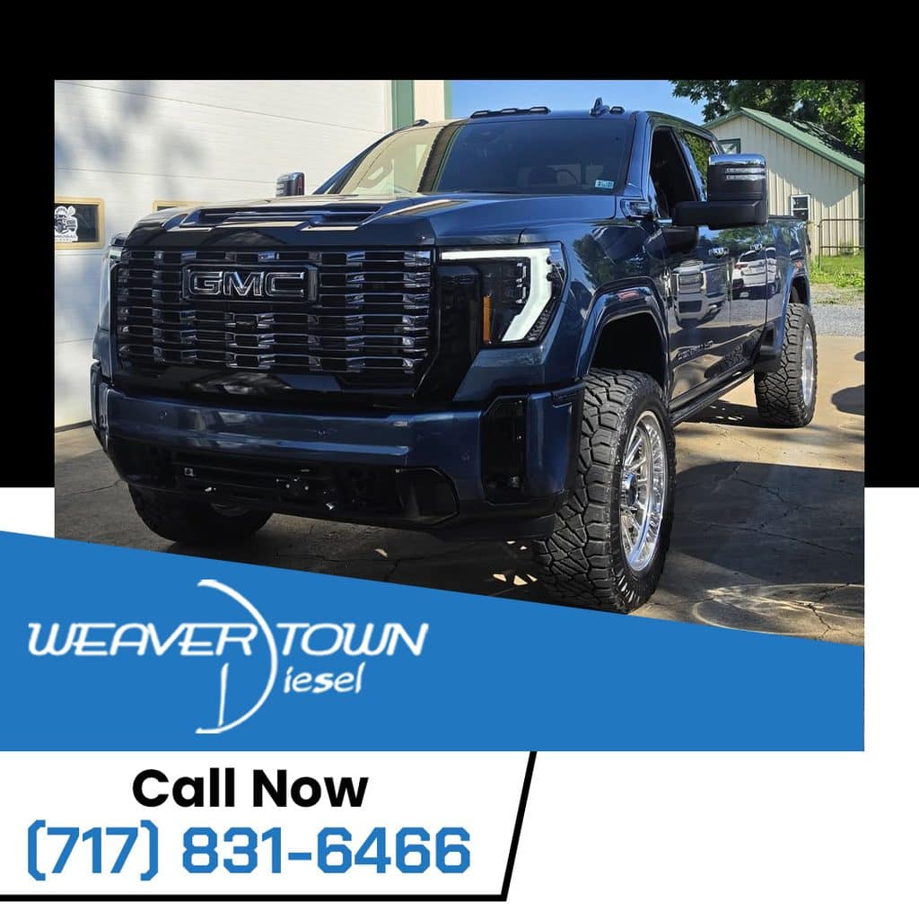 A dark blue, lifted GMC Sierra heavy-duty diesel truck equipped with aftermarket chrome wheels and rugged off-road tires parked outside a Weavertown Diesel shop facility.